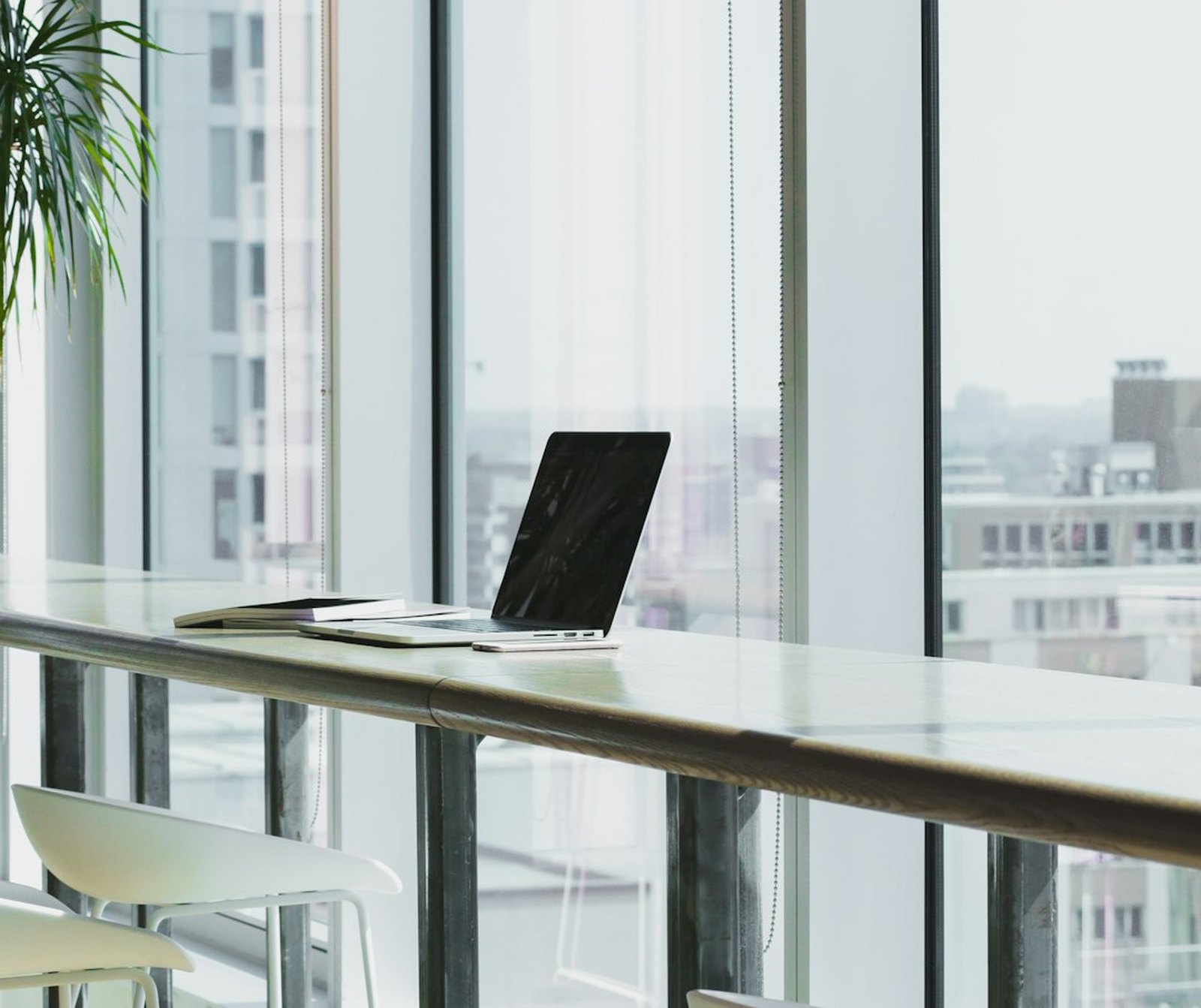 A laptop open on a work surface beside tall office windows